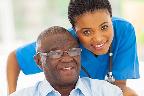 Young nurse in blue scrubs smiling behind elderly man in wheelchair wearing glasses